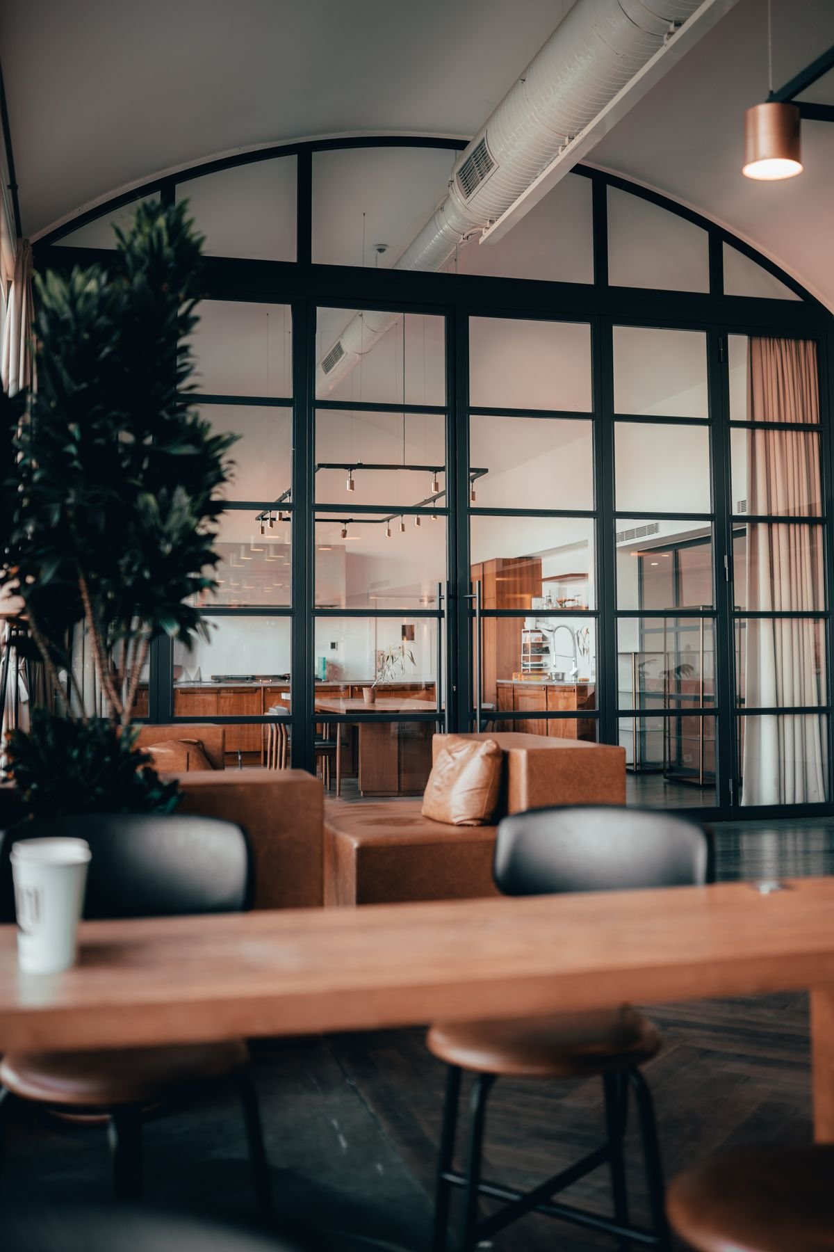A timber table and tan leather seating beneath a tall arched steel-framed window