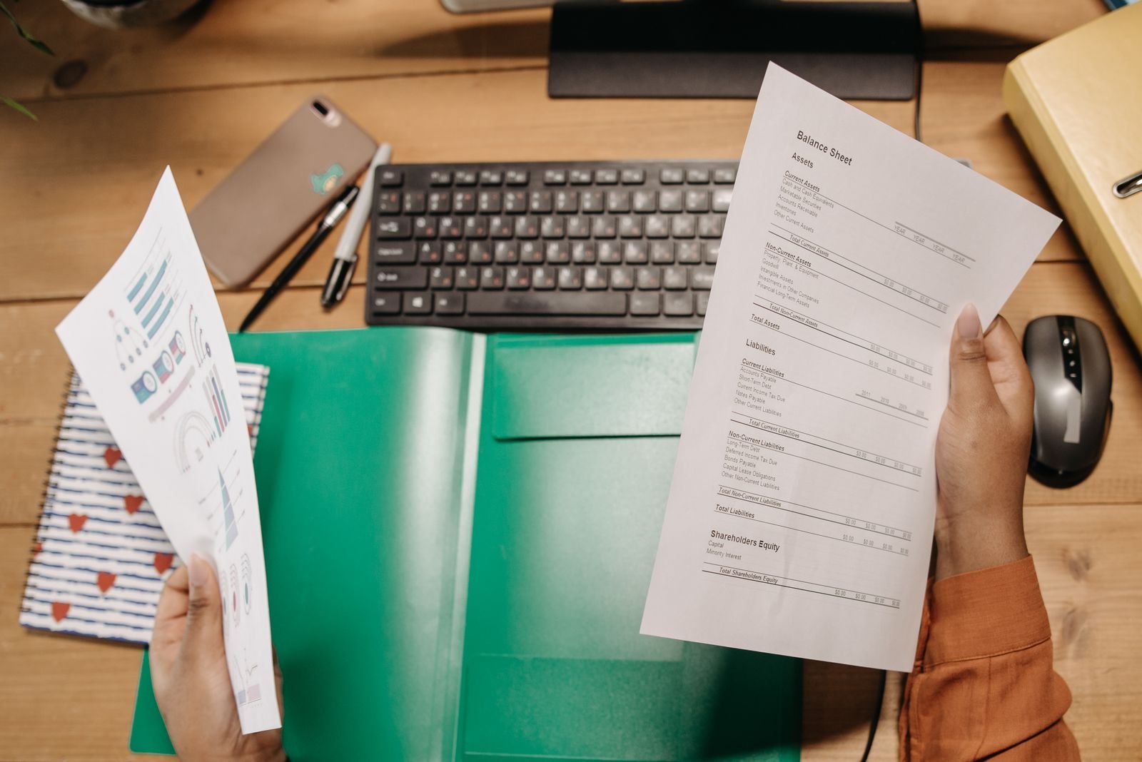 A balance sheet and a printed chart held above a green document folder on a timber desk