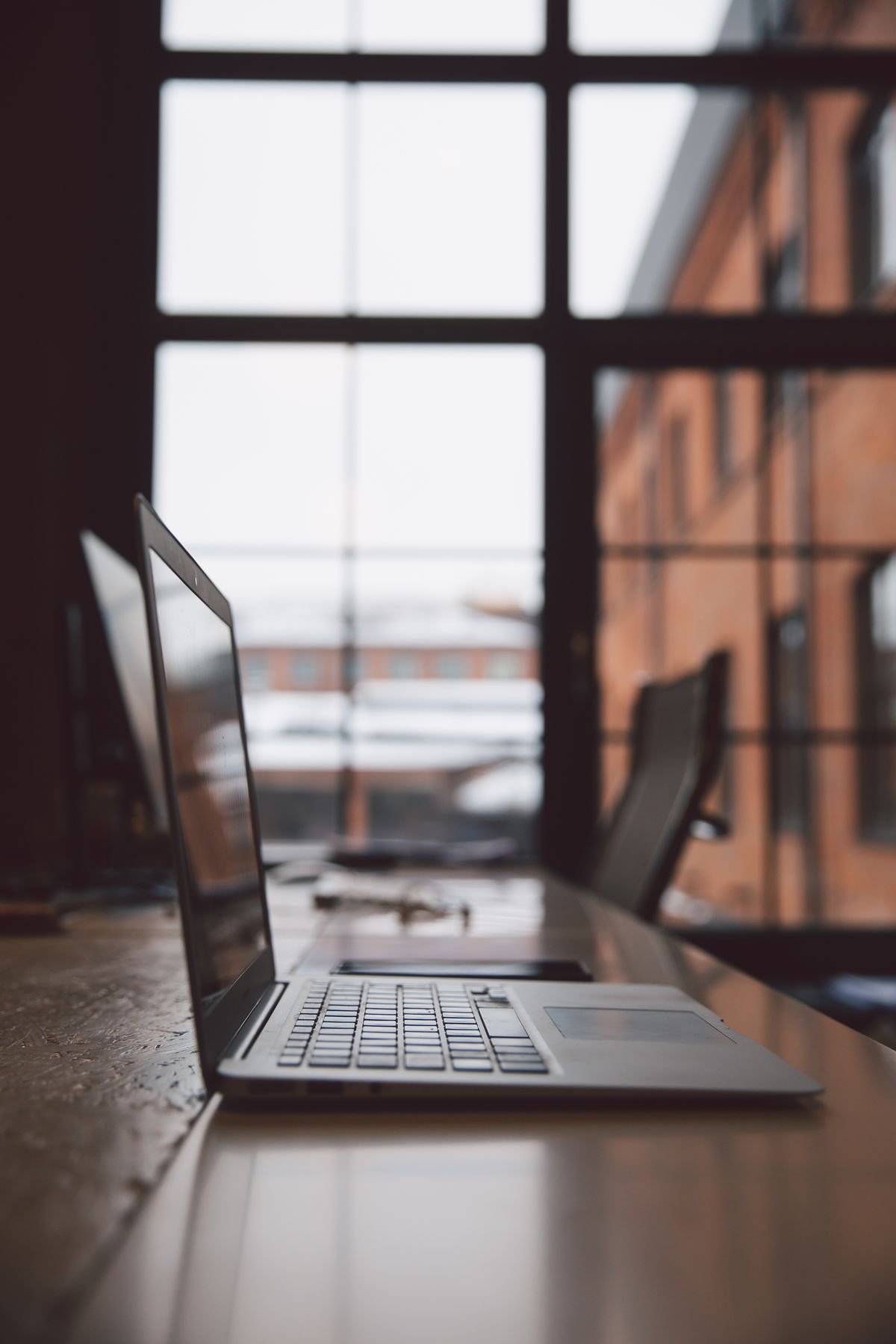 A laptop open on a long timber bench beside tall factory windows